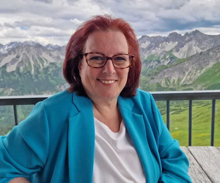 Portrait Claudia Schuster Claudia Schuster sitzt in den Bergen, lächelnd vor einer Aussicht.